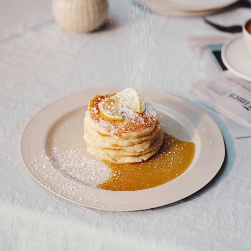 The image shows a plate of pancakes topped with powdered sugar and a lemon slice, with a cup of coffee, a newspaper, and tableware in the background.