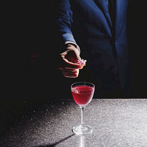 A hand in a suit jacket adds a small garnish to a red cocktail in a stemmed glass, on a reflective surface with a dark background.