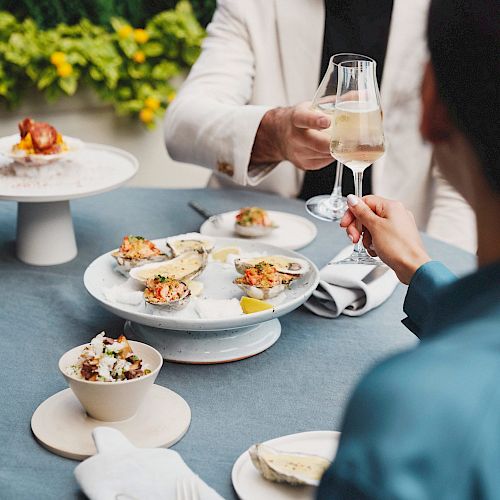 Two people are dining outdoors, enjoying a meal with various dishes including oysters. They are toasting with glasses, creating a celebratory atmosphere.