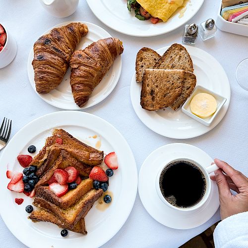 A breakfast spread with croissants, toast, an omelet with veggies, French toast with berries, a cup of coffee, and condiments on a white tablecloth.