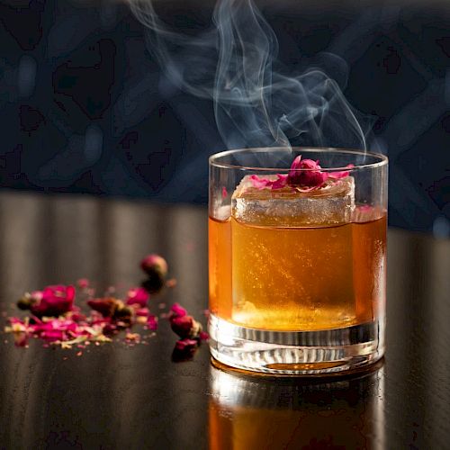 A glass containing a smoky cocktail with a large ice cube and garnished with flower petals, placed on a dark, reflective surface.