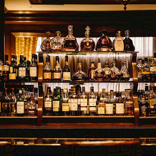 A well-stocked bar shelf filled with various types of liquor bottles, including whisky, tequila, rum, and more, arranged neatly in a wooden cabinet.