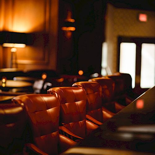 A cozy, dimly-lit bar interior with leather seating and warm ambient lighting, featuring a row of bar stools and elegant wood paneling.