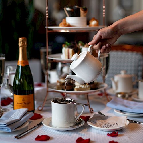 A table set for afternoon tea with a cake stand, pouring tea, bottle of champagne, scattered rose petals, and place settings.