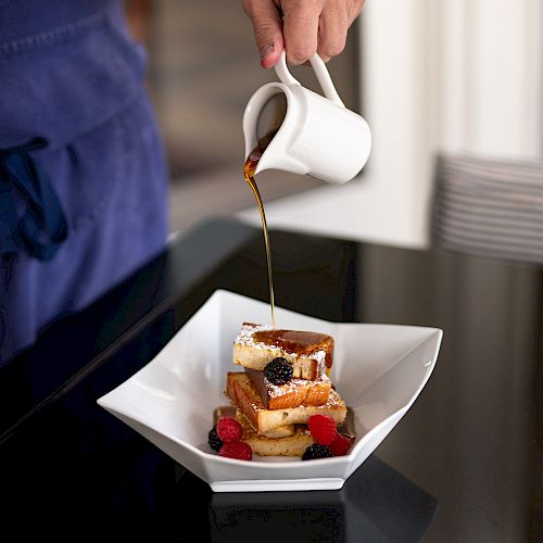 A person is pouring syrup from a white pitcher onto a stack of French toast topped with berries and powdered sugar on a square white plate.