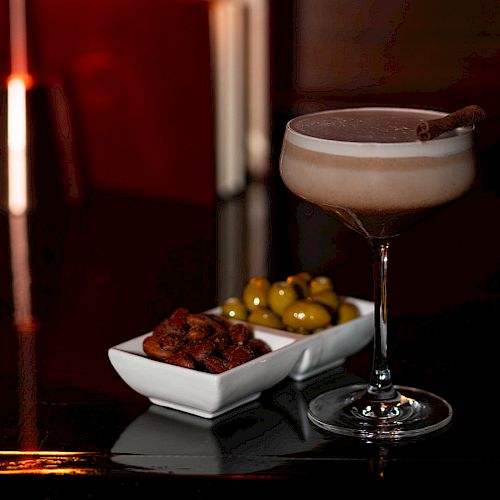 The image shows a cocktail in a glass garnished with a cinnamon stick, next to a dish of green olives and dried tomatoes on a dark surface.