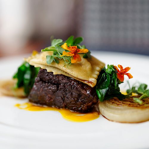 A gourmet dish featuring a piece of meat topped with vegetables, herbs, and edible flowers, served with a grilled onion slice on a white plate.