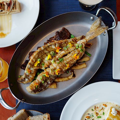 An assortment of dishes including a whole fried fish, a cocktail, a dessert, and various plated items on a wooden table is shown.