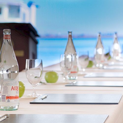 A conference table is set with bottles of water, glasses, green apples, notepads, and pens, in front of a podium and large windows with a water view.