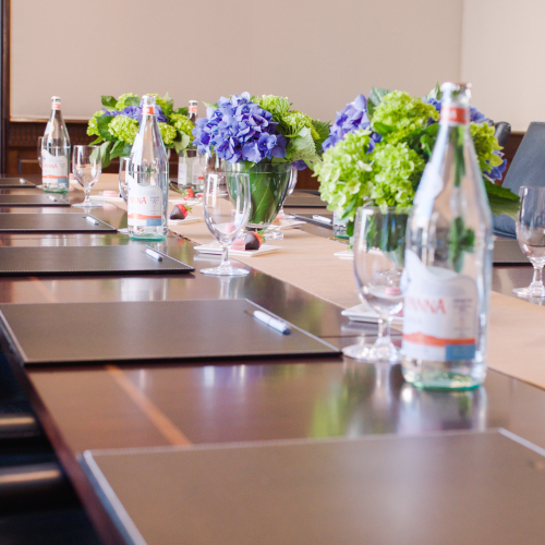 A conference room setup with black chairs, notepads, pens, water bottles, glasses, and floral arrangements in the center.