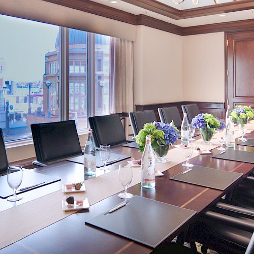 A well-lit conference room with a long table, black chairs, water bottles, glasses, notepads, pens, and flower arrangements, facing a large window.