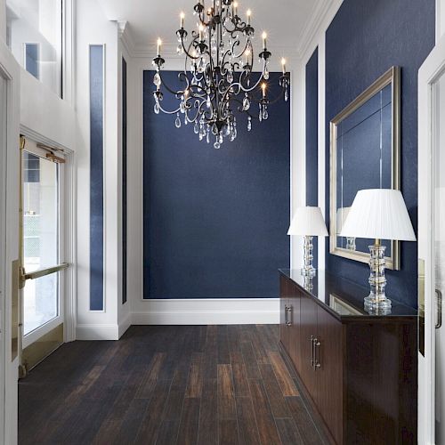 The image shows an elegant hallway with dark blue walls, a chandelier, a side table with two lamps, a large mirror, and glass doors.