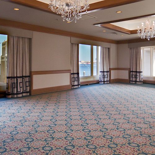 This image shows an empty room with large windows, chandeliers, and patterned carpeting, offering a view of boats and a building outside.