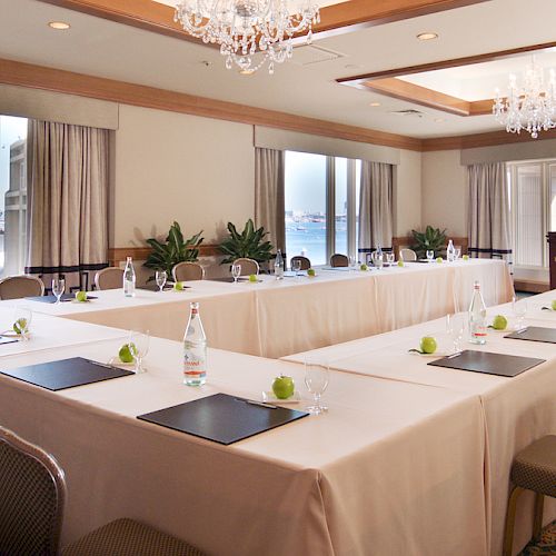 A conference room with U-shaped table setup, featuring notepads, water bottles, and green apples, large windows offer a view of a waterfront.