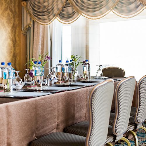 A lavish dining area with upholstered chairs, a bar setup with water bottles and glasses, and ornate drapes in front of a large window.