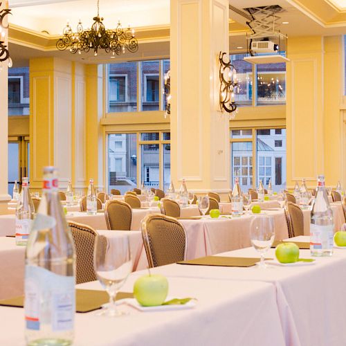 A conference room is set up with rows of tables, each featuring bottled water, apples, glasses, and notepads, with chandeliers and large windows.