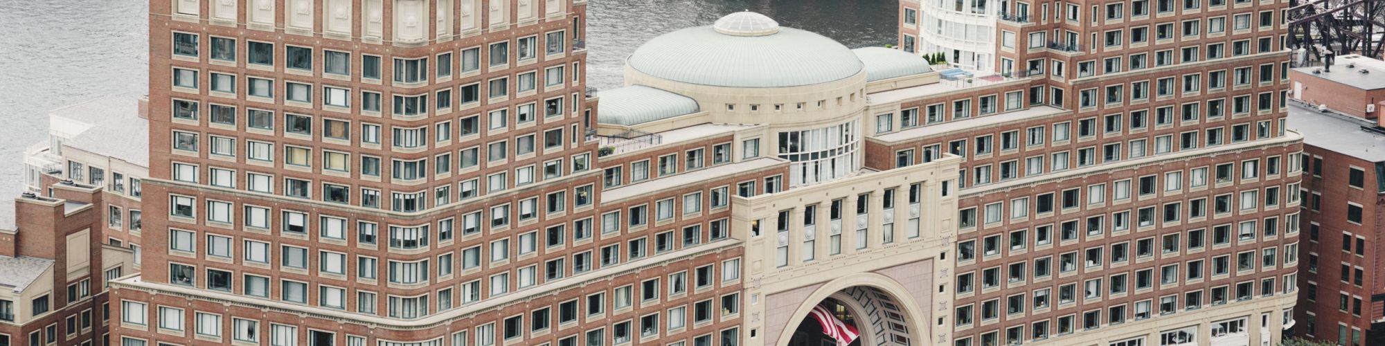 An aerial view of a large building complex with a central archway, surrounded by greenery and situated near a waterfront.