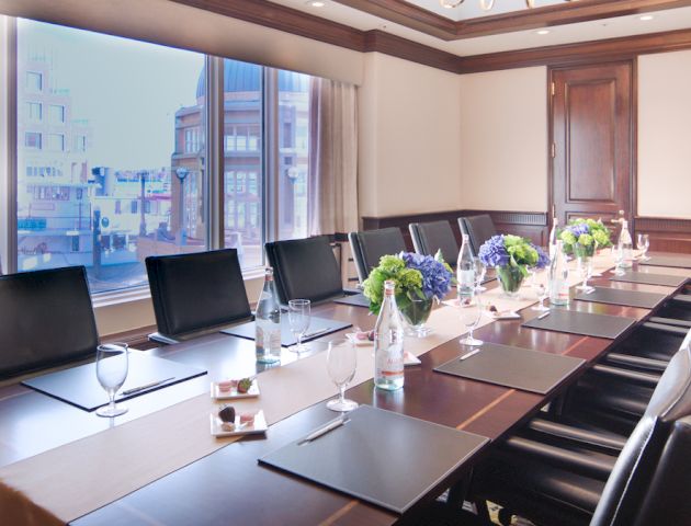 A modern conference room with a long table, black chairs, notepads, bottles of water, glasses, and floral arrangements, featuring a large window.