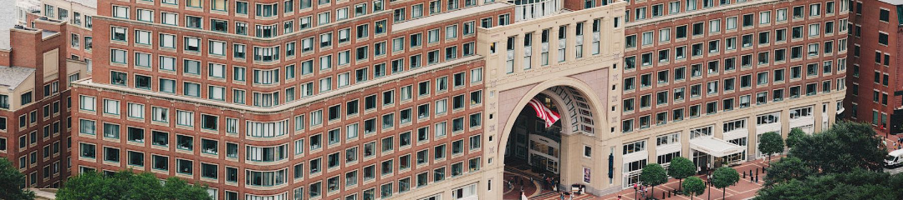 The image shows an archway building by the waterfront with people walking around and trees surrounding it.