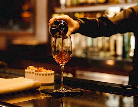 A person is pouring red wine into a glass at a bar, with snacks and napkins placed on the counter.