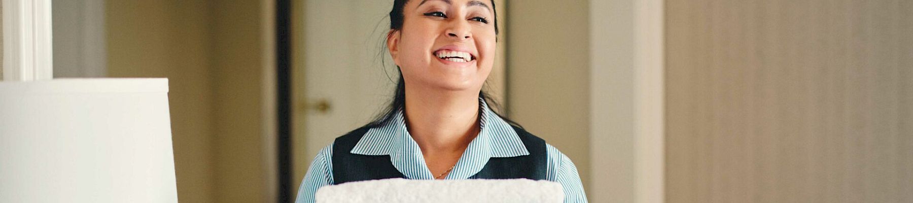 A person, smiling, is holding a stack of white towels in what seems to be a hotel room with beige walls and a white lamp.