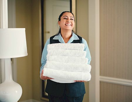 A person, smiling, is holding a stack of white towels in what seems to be a hotel room with beige walls and a white lamp.