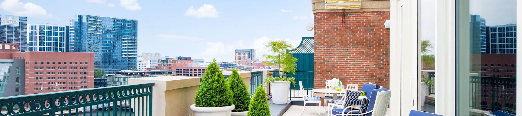 A modern rooftop terrace with blue cushioned seating, plants, and a view of city buildings and a body of water, under a clear blue sky.