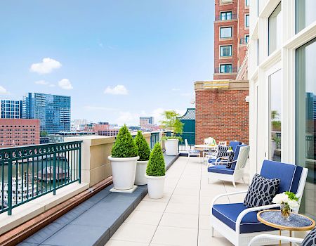 A modern rooftop terrace with blue cushioned seating, plants, and a view of city buildings and a body of water, under a clear blue sky.