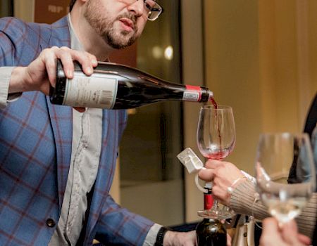 A man is pouring red wine into a glass held by another person. Other wine bottles are visible in the foreground, along with framed artwork.