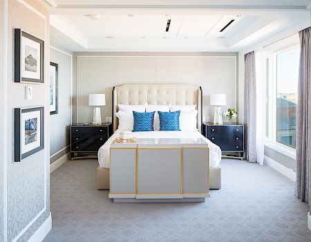 This is a well-decorated bedroom featuring a large bed with blue pillows, nightstands, lamps, framed artwork on the walls, and a large window.