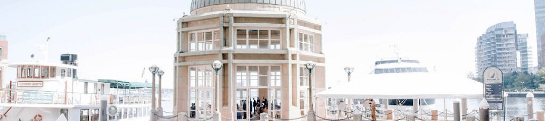 A domed pavilion by a waterfront with boats docked and city buildings in the background.