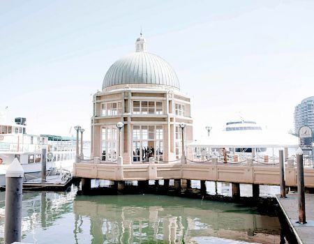 A domed pavilion by a waterfront with boats docked and city buildings in the background.