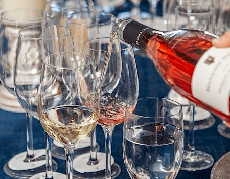 A hand is pouring rosé wine into a wine glass among various other glasses, including ones with water and white wine.