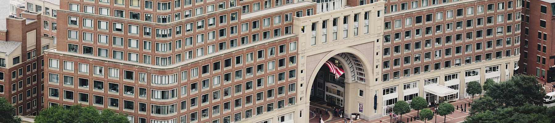 The image shows an aerial view of a large, multi-story brick building with a prominent archway near a waterfront, surrounded by urban landscaping.