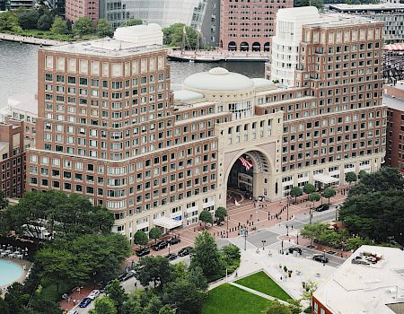 The image shows an aerial view of a large, multi-story brick building with a prominent archway near a waterfront, surrounded by urban landscaping.