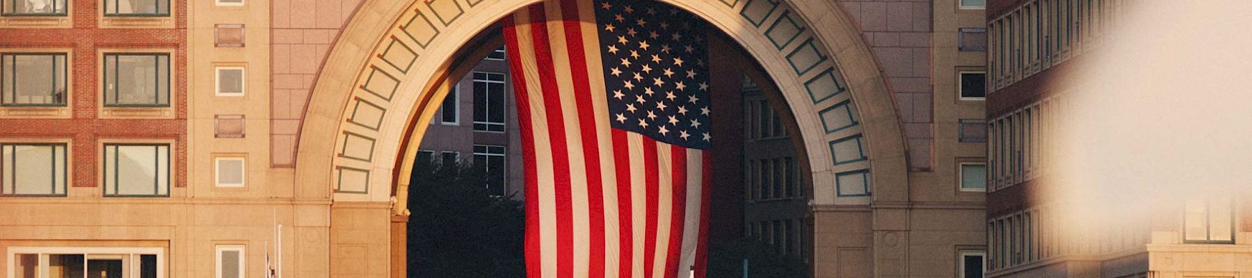 The image shows a large building with an archway displaying an American flag. There are boats docked in the water in the foreground, and city buildings.