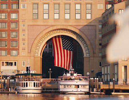 The image shows a large building with an archway displaying an American flag. There are boats docked in the water in the foreground, and city buildings.