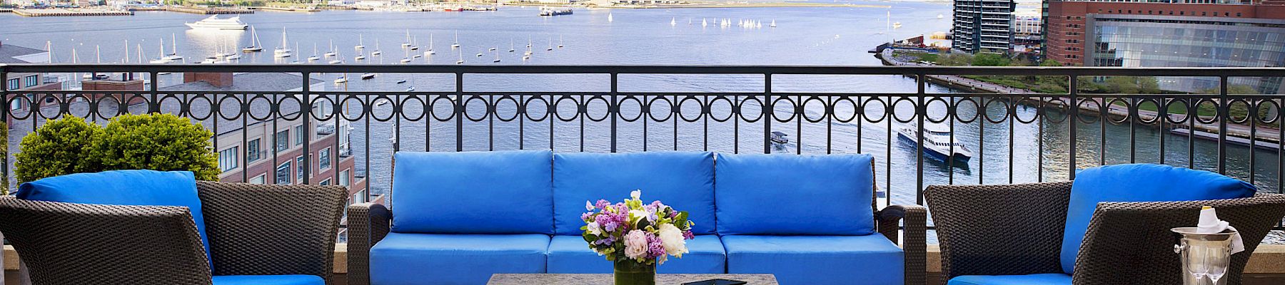 A patio with blue cushioned furniture set facing a scenic waterfront view and city buildings, with a table holding flowers between the seating.