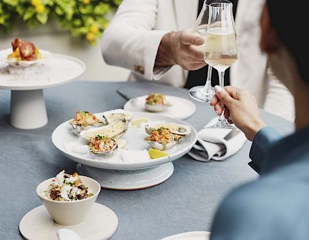 Two people are dining outdoors, clinking glasses with seafood dishes and cutlery on the table in front of them.