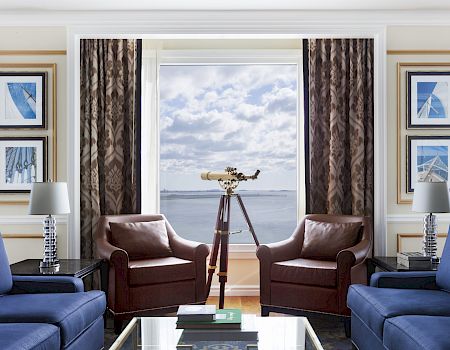 A living room with blue sofas, brown armchairs, a telescope by a large window with an ocean view, two lamps, and framed wall art.