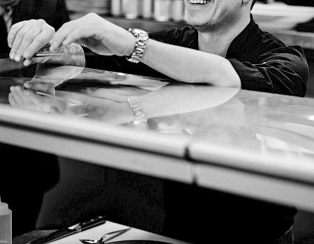 A smiling man stands behind a counter in a kitchen setting, with containers of ingredients in front of him.