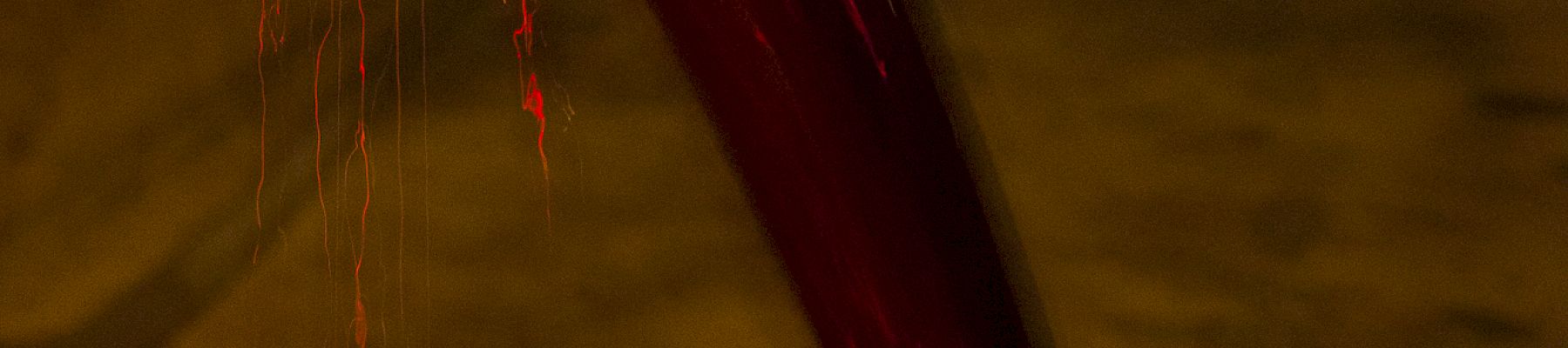 A stream of red liquid pours into a container in dim lighting, with a candle burning in the background.