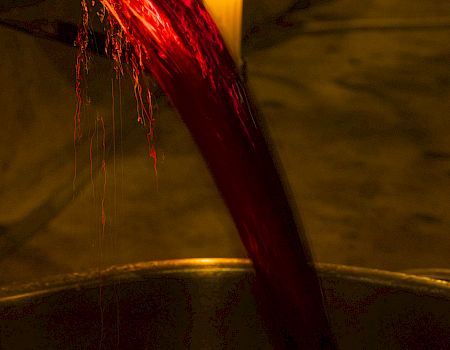 A stream of red liquid pours into a container in dim lighting, with a candle burning in the background.