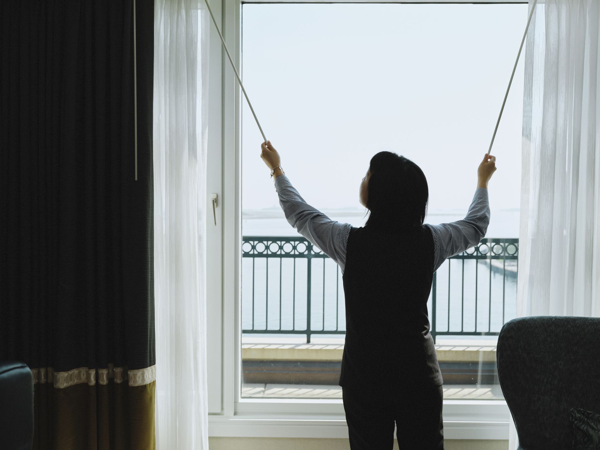 A person is standing in front of a large window, opening the curtains to reveal a view of a balcony and a body of water in the distance.