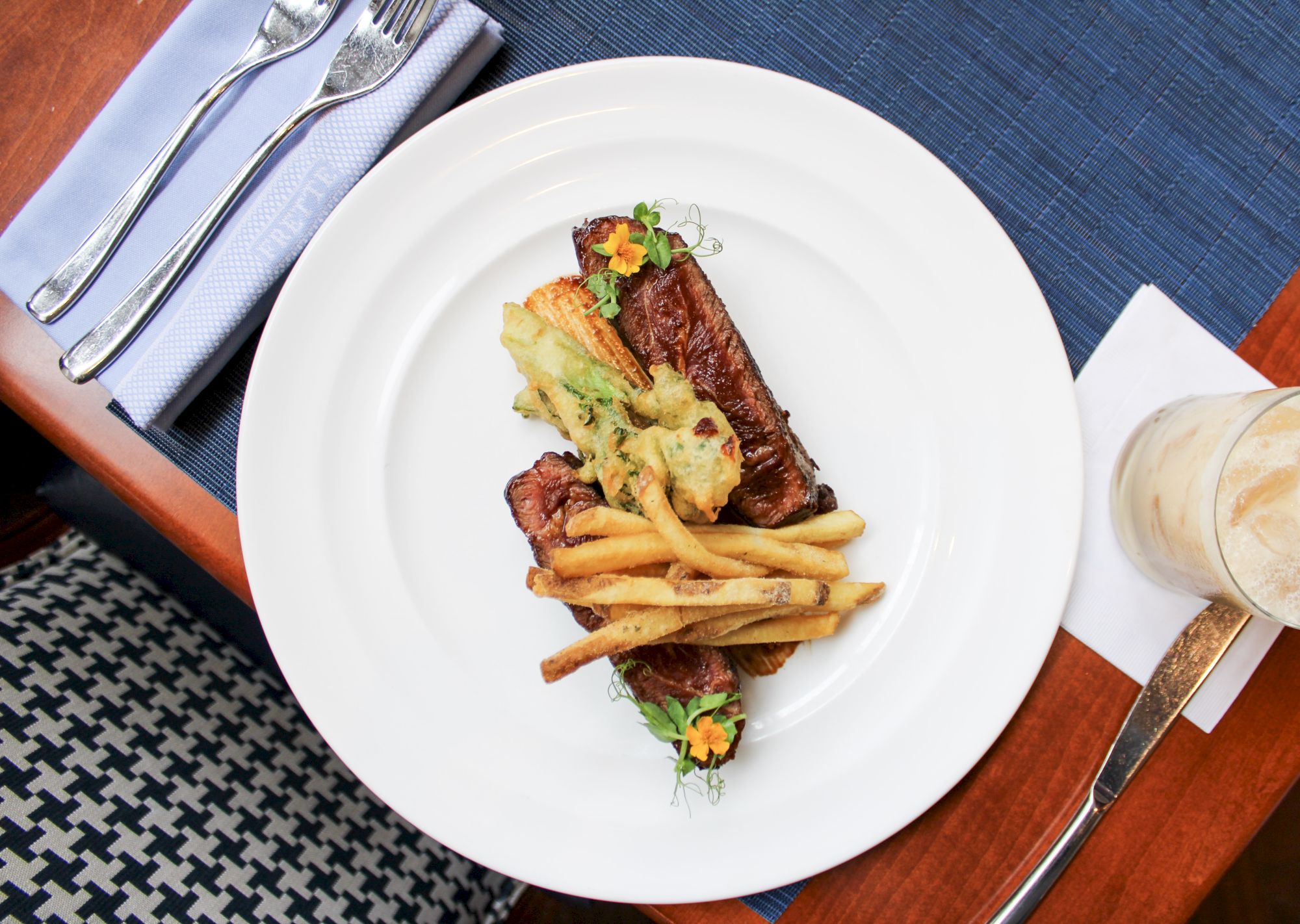 A plate with steak, fries, and vegetables, placed on a blue placemat next to a drink and utensils on a napkin, on a wooden table.