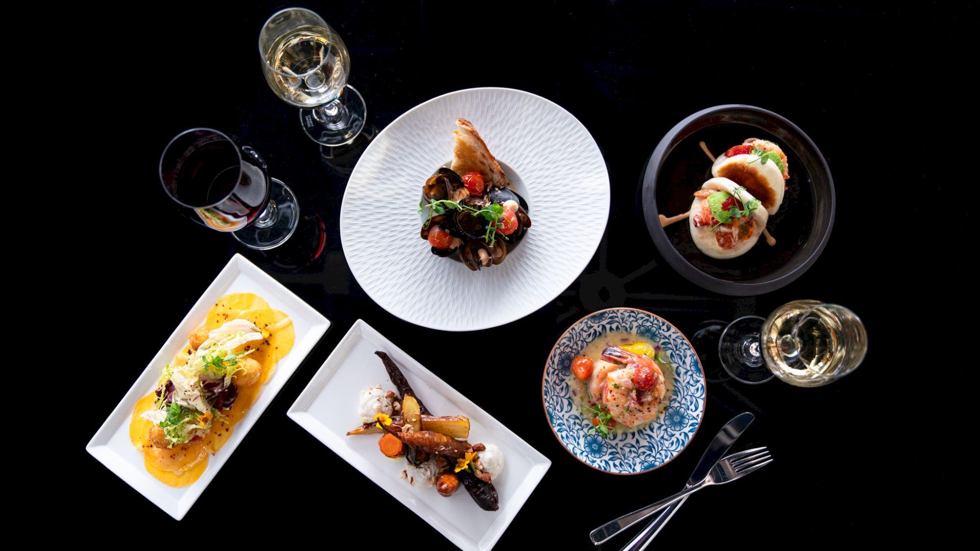 The image shows an elegant arrangement of various gourmet dishes and drinks on a black background, including wine glasses and plated meals.