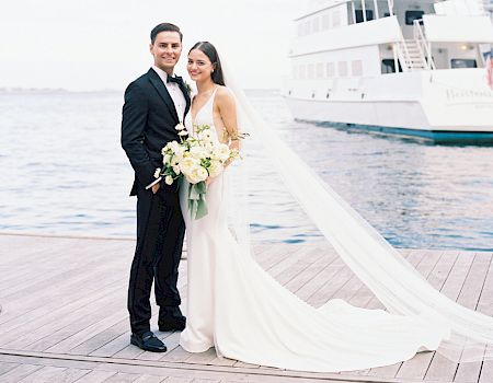 A bride and groom stand on a dock near a body of water and a large boat. The bride holds a bouquet and wears a flowing white dress with a long train.