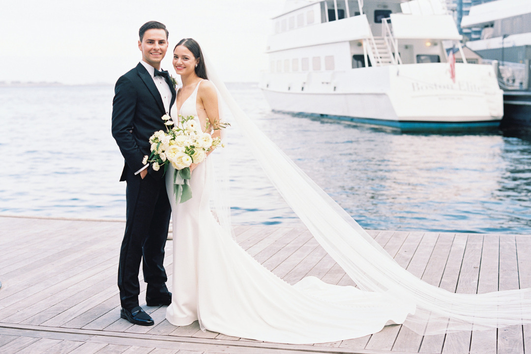 A bride and groom stand on a dock near a body of water and a large boat. The bride holds a bouquet and wears a flowing white dress with a long train.