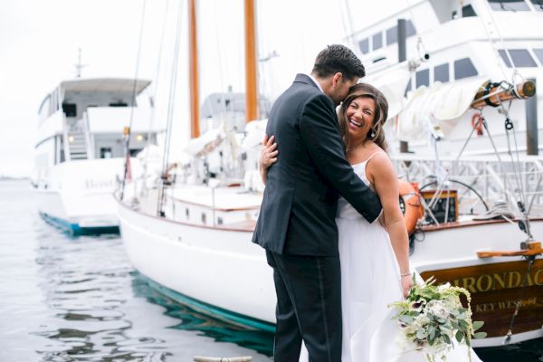 A bride and groom are smiling and embracing each other in front of boats on a dock. The bride is holding a bouquet of flowers.