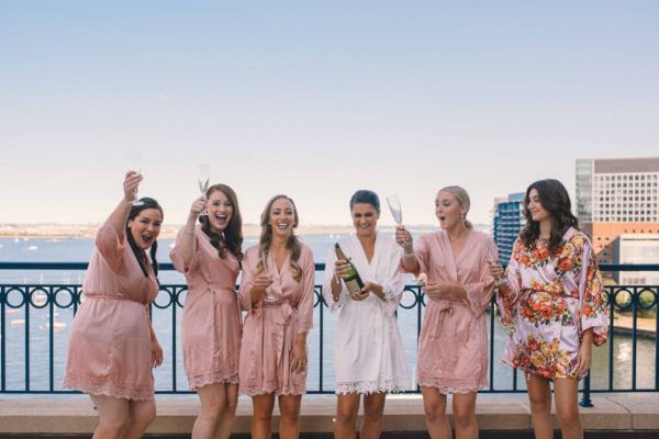 A group of women in robes celebrate with champagne on a terrace overlooking a waterfront cityscape.